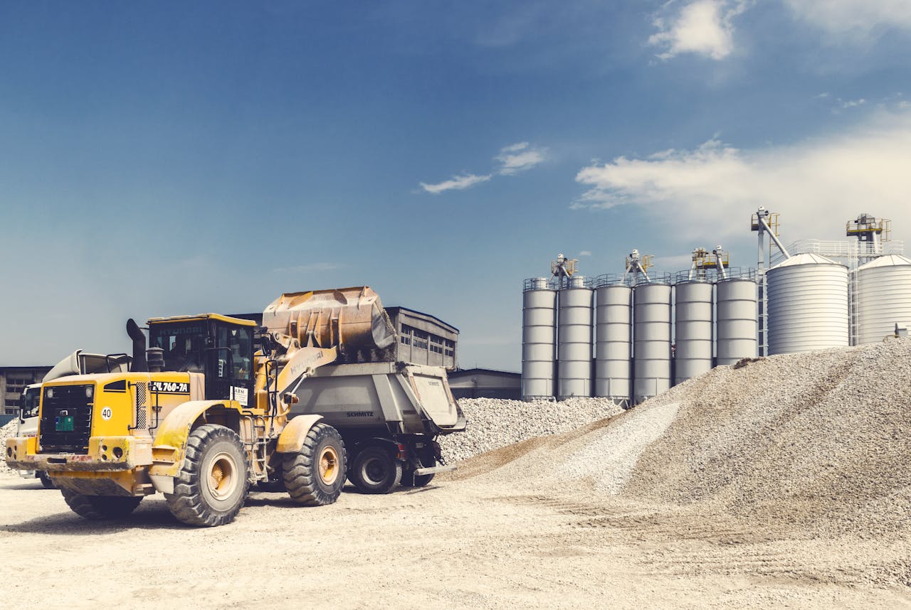 Yellow loader and truck operating at a gravel yard with silos in the background on a sunny day.
